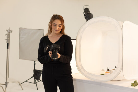 Adult woman in black clothing holding DSLR checking settings in studio with light tent and strobe