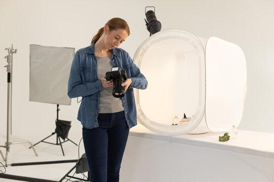 Woman in her 20s reviewing DSLR settings while leaning on table near light tent with bottles