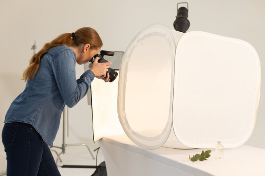 Woman in denim shirt leaning, using DSLR flash, shooting scent bottle in studio softbox, copy space