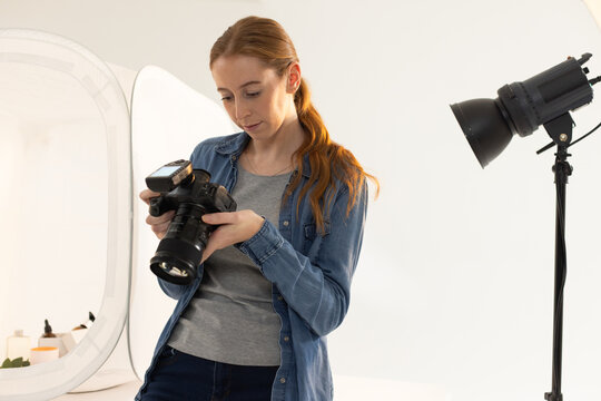 Woman in twenties wearing denim shirt holding DSLR camera and adjusting strobe beside reflector