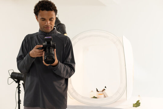 Adult African American male holding DSLR and checking controls in studio with softbox and bottles