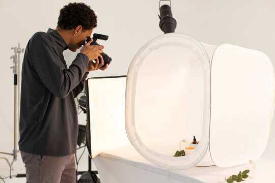 Male photographer standing at table in studio, aiming DSLR with flash into light tent, copy space