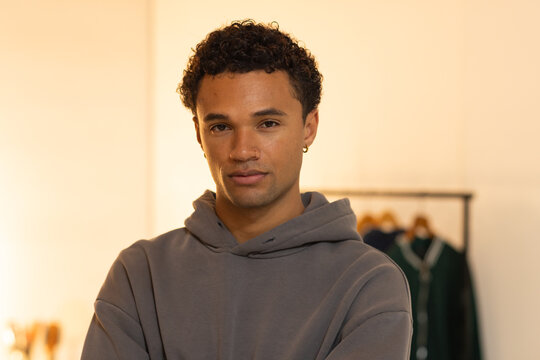 Man wearing gray hoodie and hoop earring, posing in studio with clothing rack and green garment