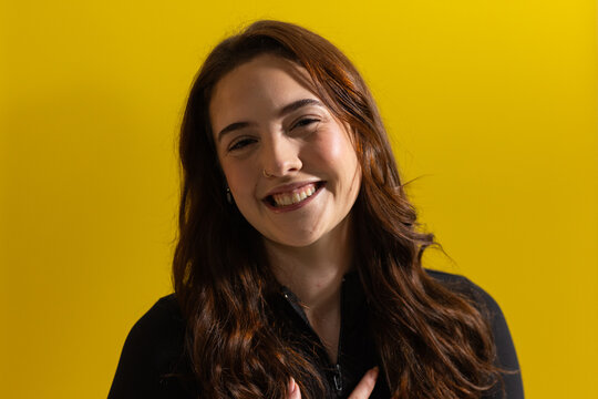 Adult woman smiling, raising hand to chest on yellow backdrop, nose ring, studs, black zip-up top