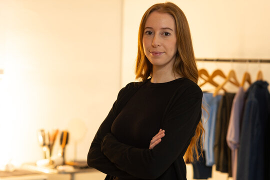 Adult female standing arms crossed, wearing black top in boutique, rack wooden hangers, table lamp