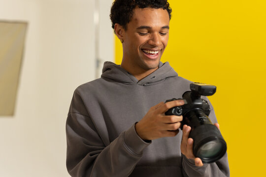 African American man 20s in gray hoodie checking DSLR with lens and flash against yellow backdrop