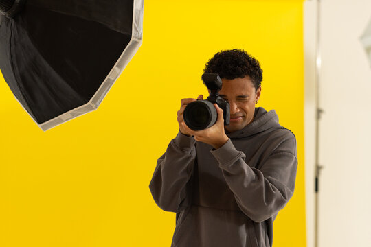 Man in hoodie holding DSLR with flash, aiming through finder on yellow studio backdrop near softbox