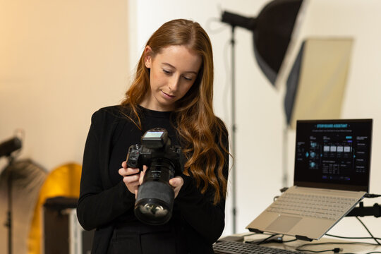 Woman in 20s-30s holding DSLR with telephoto lens and checking camera display with laptop in studio