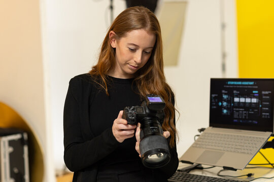 Woman in her 20s wearing black holding DSLR and adjusting settings in studio with laptop, reflector