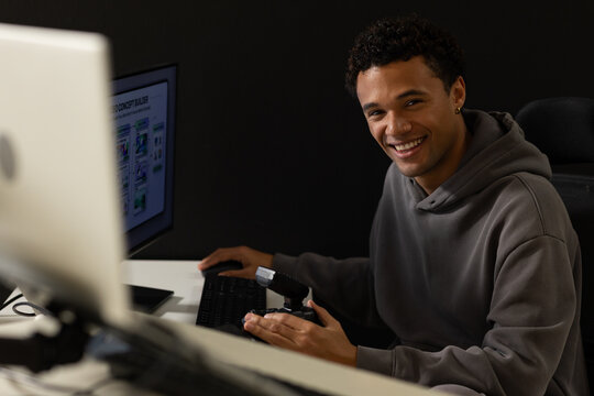 African American man in hoodie smiling while adjusting flash at desk with dual monitors, copy space