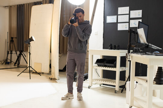 African American male holding DSLR camera and framing shot in photography studio with lenses and PC
