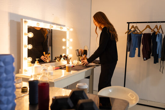 Female stylist arranging makeup brushes on dark mat at lit vanity mirror, clothing rack, copy space