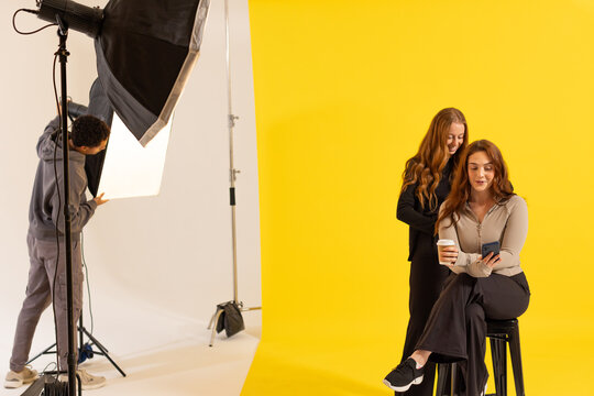 Two female models holding cup and phone, one looking while male tech adjusting softbox in studio
