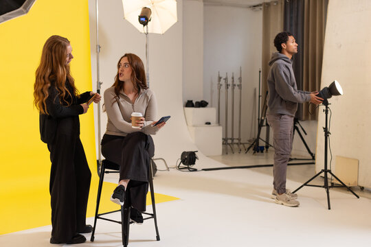 Diverse crew collaborating in studio, adjusting lights, model on stool holding coffee and phone
