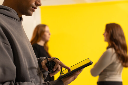 Male tech in hoodie using tablet, remote on set, models posing on yellow backdrop, copy space