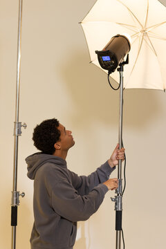 African American man adjusting strobe and white umbrella on light stand in gray hoodie, copy space