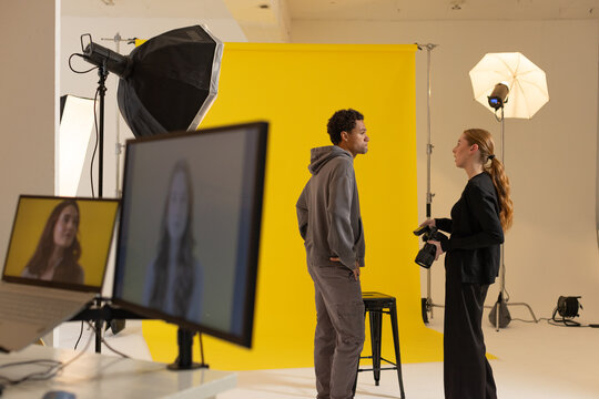 Male model posing by yellow studio backdrop, photographer in black holding DSLR screen showing face
