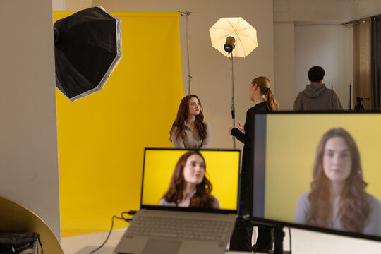 Studio team adjusting lights while shooting seated model on yellow backdrop, watching laptop
