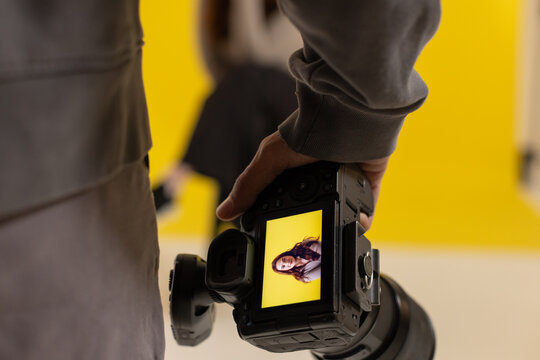 Male photographer holding DSLR, checking LCD against yellow backdrop, female model sitting on stool