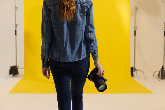 Female photographer walking toward yellow backdrop in studio holding DSLR and wearing denim shirt