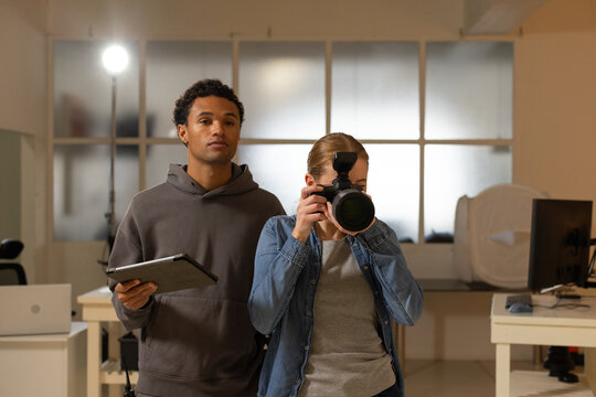 Diverse coworkers shooting in studio, photographer using DSLR flash, assistant holding tablet