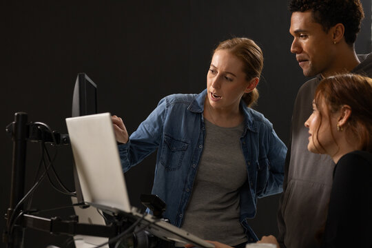 Diverse coworkers leaning and pointing at laptop on monitor mount with cables and camera in studio