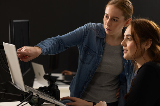 Two female colleagues collaborating around desk workstation pointing at laptop using mouse and DSLR