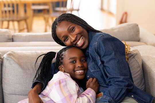 African mother and youth daughter hugging and smiling on gray sofa wearing denim and pastel-plaid