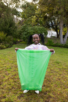 African youth female standing on lawn, holding green plastic bag and wearing white sneakers
