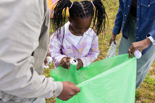 African family collecting litter on grassy park, youth placing aluminum can into green trash bag