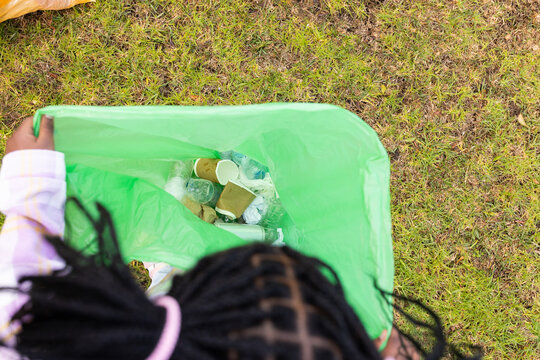 African youth female holding open green trash bag on patchy lawn, gathering cups, bottles, sleeves