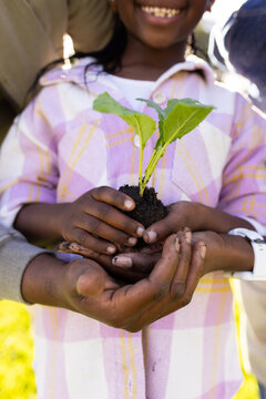 Family supporting child in park wearing pink plaid shirt holding seedling with soil and white watch