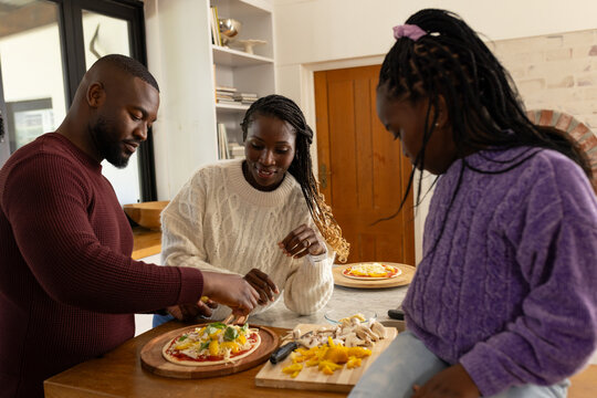 African American family in sweaters assembling pizzas on wooden kitchen island, using pizza board