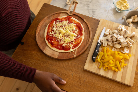 African American man in maroon sweater preparing pizza on wooden peel in kitchen, knife, mushrooms