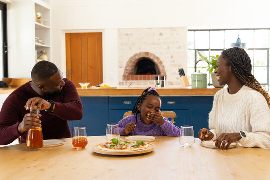 African American family sharing pizza at kitchen table, father pouring pitcher, child laughing