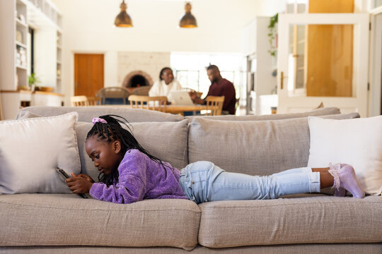 African family sitting at dining table working on laptop while child lying on sofa using smartphone