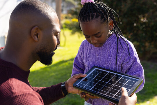 African American man holding solar panel while child inspecting on yard, in maroon and purple knits
