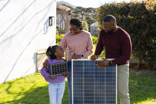 African American family standing in backyard by house holding large and small solar panels