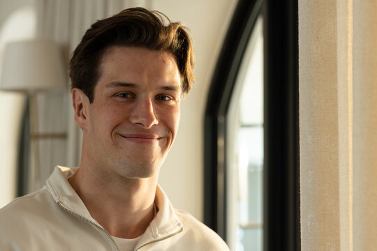 Man standing near arched black-framed window at home, wearing light zip-up top smiling