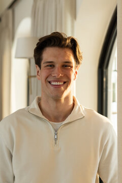 Man standing smiling facing camera at home wearing cream pullover near arched window and lamp