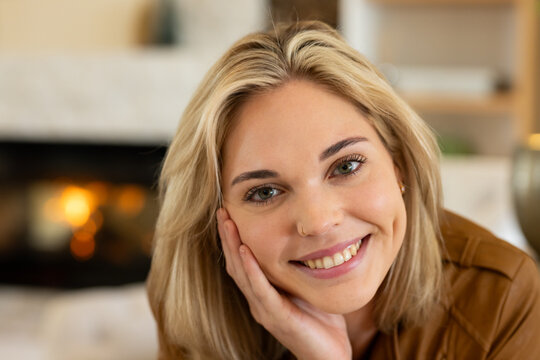Adult female smiling, leaning head on hand, wearing brown jacket, showing nose ring by fireplace