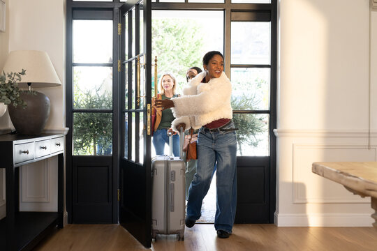 Three female friends entering home entryway pulling silver suitcase and carrying brown shoulder bag