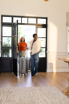 African women entering foyer through black-framed glass door, pulling silver suitcase, holding door