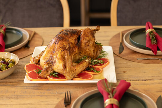 Roasted whole turkey is resting on white platter on wooden table, garnishing with citrus, rosemary