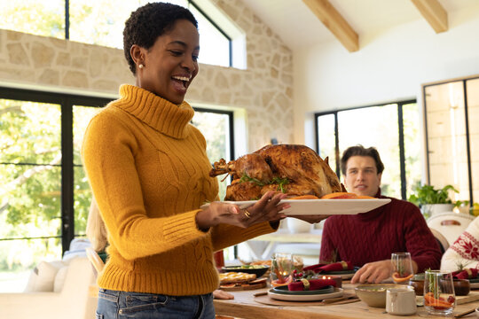 African American woman in mustard knit holding turkey on white tray in dining room, copy space