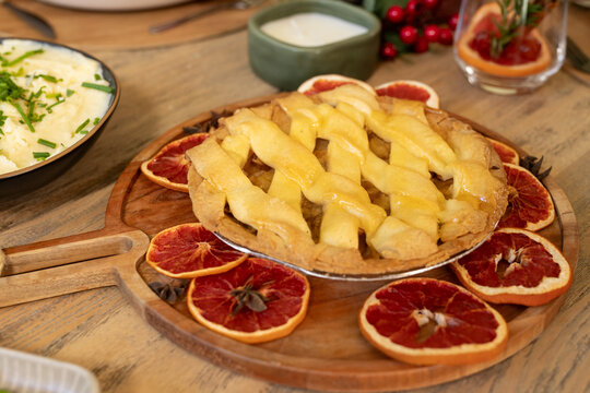 Lattice fruit pie is resting on wooden serving board on table beside mashed potatoes, dried citrus