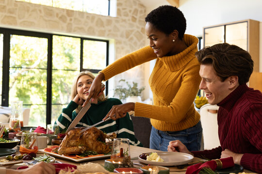 Diverse friends watching African American woman carving turkey with knife in mustard top at table
