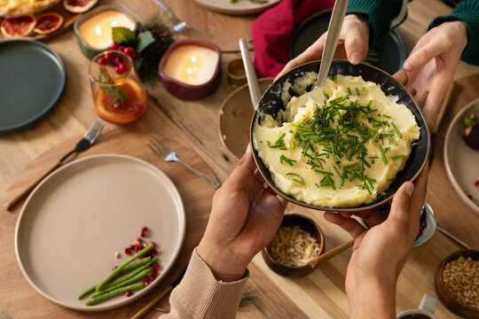 Bowl of mashed potatoes being passed across wooden dining table during festive meal with chives