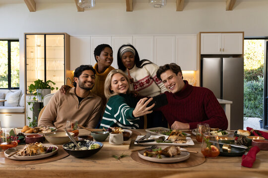 Diverse friends taking selfie at wooden dining table with phone, plates of food, wearing sweaters