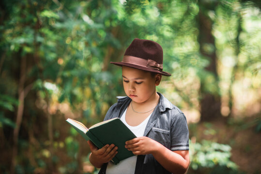 Boy reading an open book outdoors in a forest. Thoughtful little boy wearing a hat holding and reading an open book outdoors in a scenic green forest.